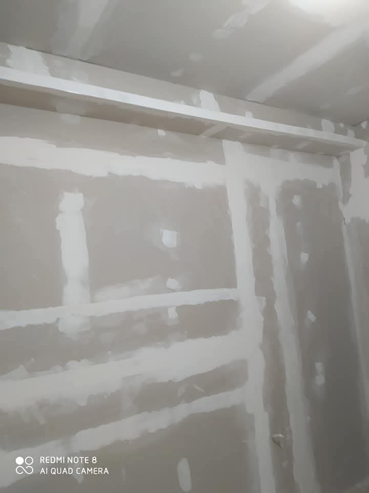 Άβαφη γυψοσανίδα με ορατές κολλημένες με ταινία και λασπωμένες ραφές σε εξέλιξη κάτω από το φωτιστικό οροφής. Ένα λευκό ράφι εκτείνεται κατά μήκος του πάνω μέρους του τοίχου.
