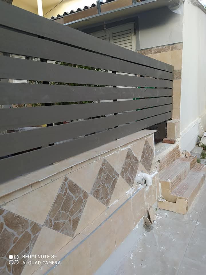 Ένας καφέ οριζόντιος φράχτης από σχιστόλιθο βρίσκεται στην κορυφή ενός πλακόστρωτου τοίχου, δίπλα σε σκαλοπάτια που οδηγούν σε μια πόρτα.