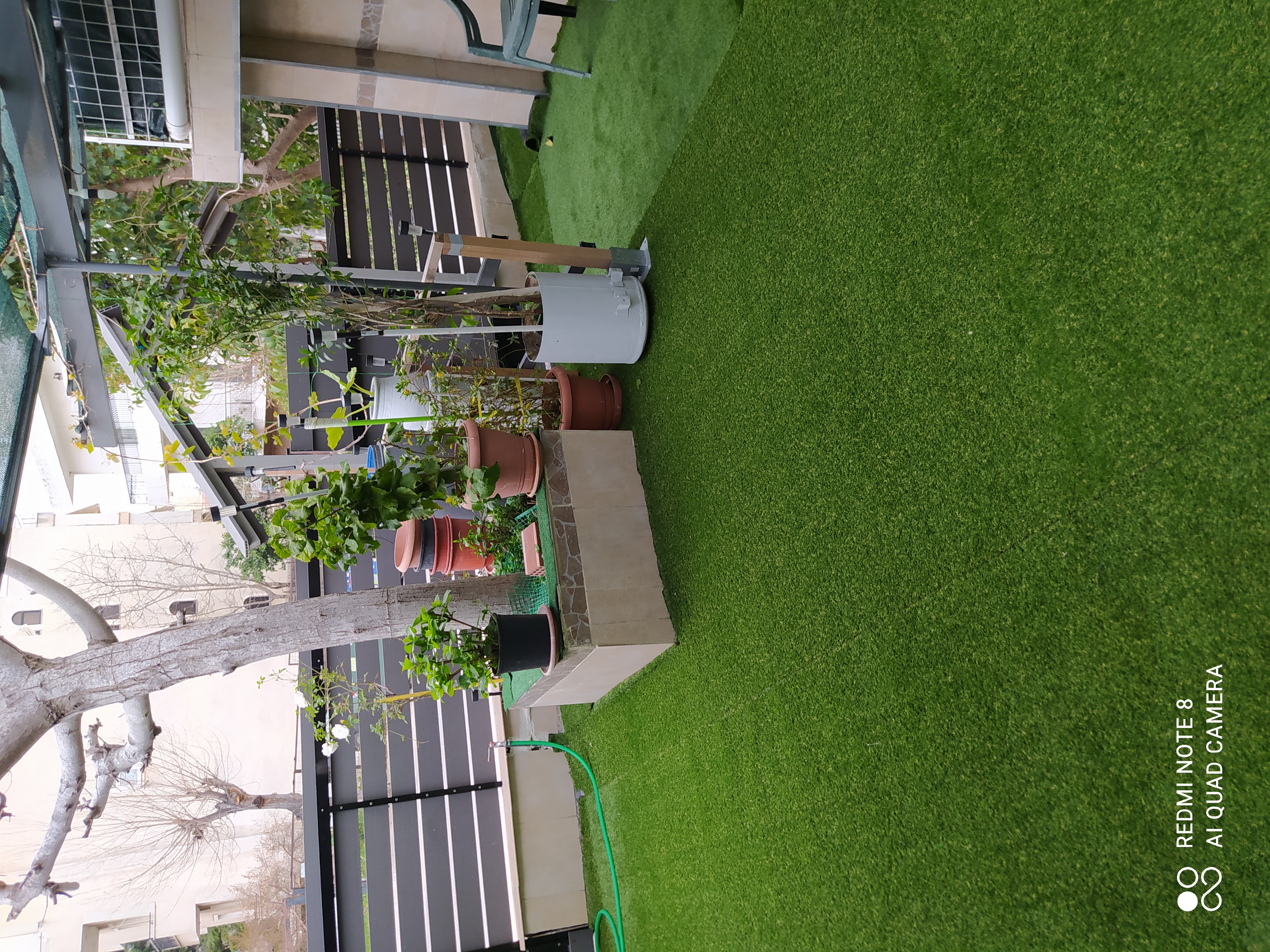 Μια βεράντα με τεχνητό γρασίδι, φυτά σε γλάστρες, μια υπερυψωμένη ζαρντινιέρα, ένα δέντρο, ένα λάστιχο νερού και έπιπλα εξωτερικού χώρου δίπλα σε ένα περιφραγμένο όριο.